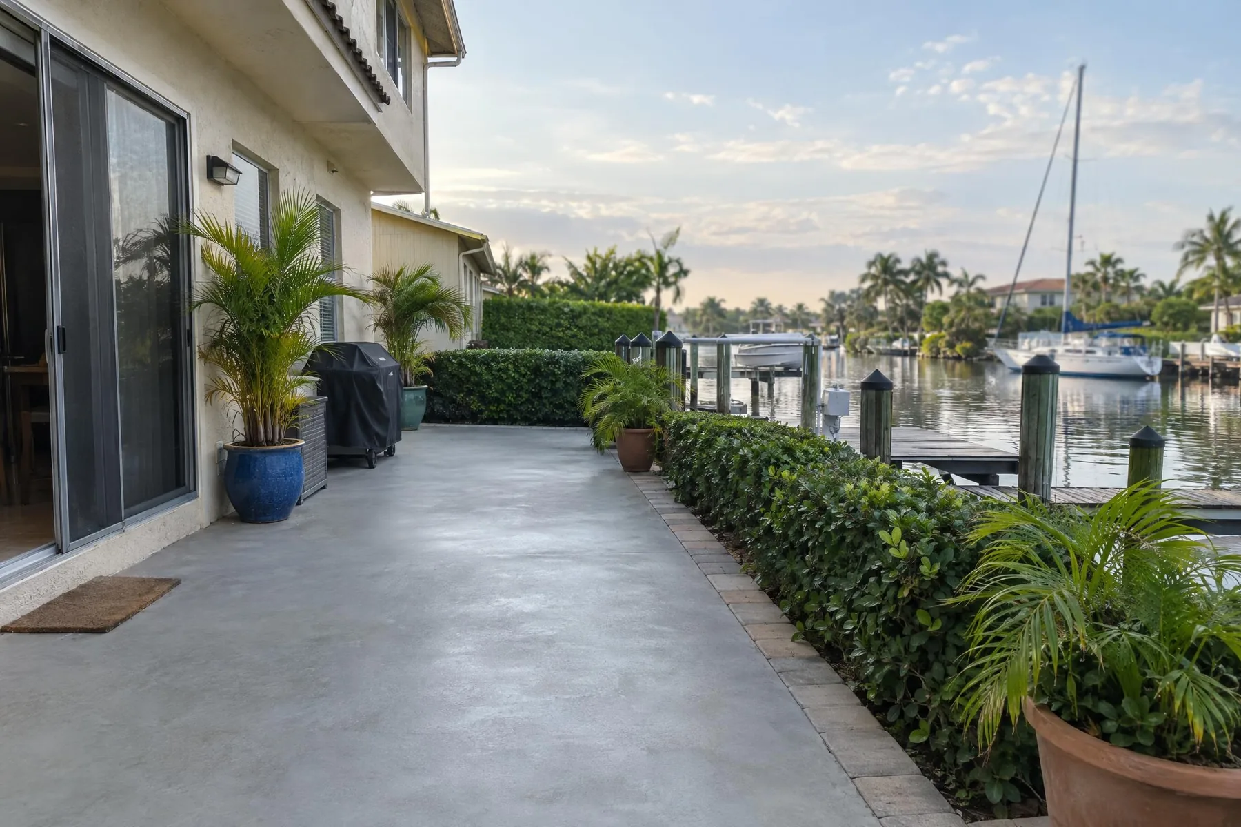 A concrete patio running along a canal with a boat dock and a trimmed hedge beside it